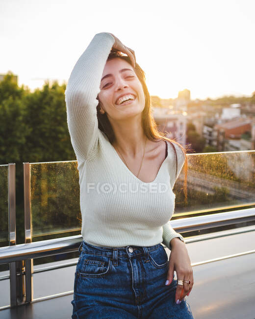 Fröhliche Frau mit braunen Haaren in lässiger Kleidung sitzt am Holztisch und lacht im Sonnenlicht auf der Straße — Stockfoto