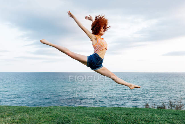 Pieno corpo di femmina scalza facendo scissione mentre salta in alto con le braccia sollevate sulla costa del mare increspato — Foto stock