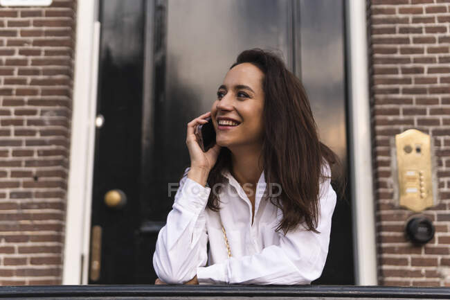 Vue latérale d'une jeune femme heureuse en vêtements décontractés debout près de l'entrée du bâtiment et appuyée sur la main courante tout en faisant un appel téléphonique — Photo de stock