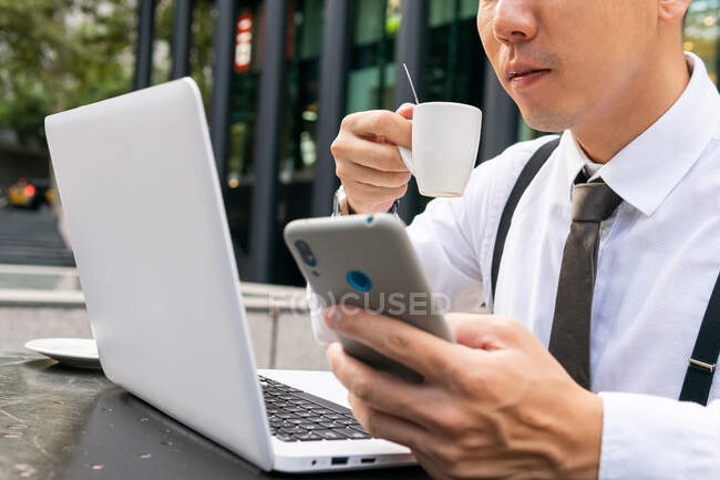 Cropped unrecognizable ethnic male entrepreneur with coffee surfing internet on cellphone while sitting at urban cafeteria table with laptop — Stock Photo