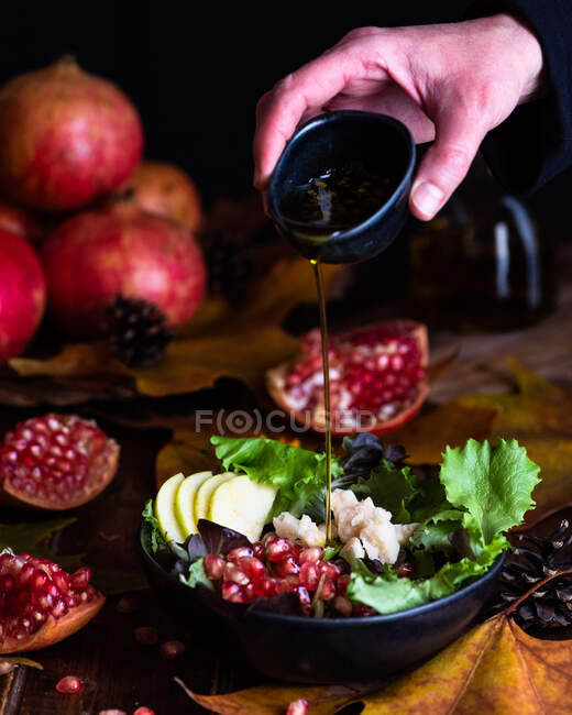 Crop chef anónimo añadiendo aceite de oliva en un tazón con apetitosa ensalada de granada vegetariana servida en la mesa con hojas y conos de otoño - foto de stock
