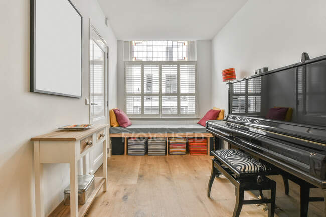 Habitación moderna vacía con paredes blancas y suelo de madera con piano negro y sofá decorado con cojines de colores y cajas de almacenamiento durante el día - foto de stock