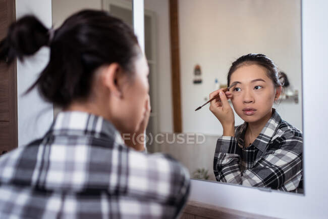 Focada encantadora fêmea étnica aplicando lápis de sobrancelha enquanto faz maquiagem e olhando no espelho — Fotografia de Stock