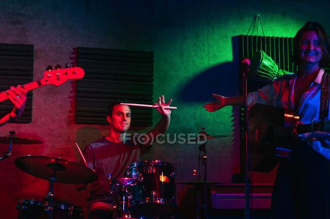 Giovani che eseguono musica con batteria e chitarre vicino al microfono in club con illuminazione al neon mentre la donna introduce il batterista — Foto stock