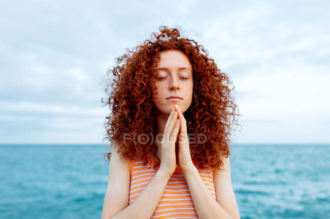 Mulher pacífica de pé com os olhos fechados na praia e fazendo gesto namaste durante a meditação — Fotografia de Stock