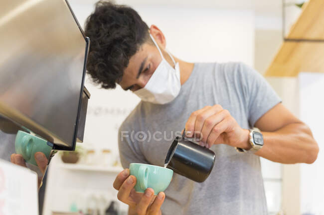 Barista étnico en máscara protectora verter leche en taza con café mientras está de pie en el mostrador con cafetera y vajilla en la cafetería - foto de stock
