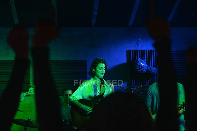 Groupe de personnes en vêtements décontractés jouant de la guitare et de la batterie pendant que la femme chante et chante une chanson en club avec des néons — Photo de stock