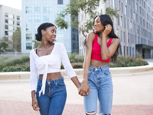 From below of optimistic African American lesbians in stylish clothes looking at each other and holding hands while walking on street with buildings — Stock Photo