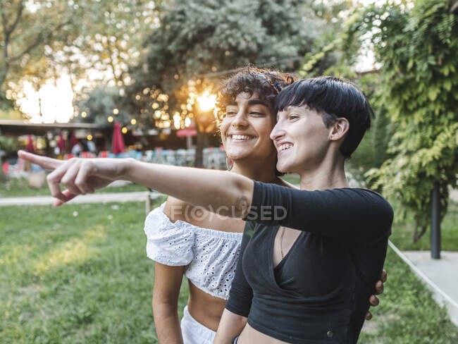 Delighted multiethnic couple of lesbian females pointing away and smiling while standing in park in summer — Stock Photo