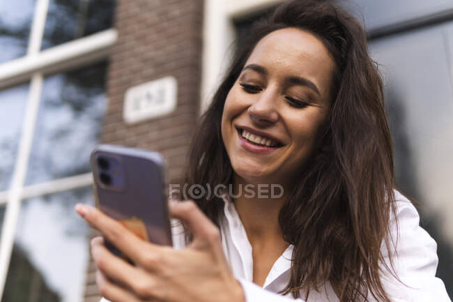 Крупный план счастливой молодой женщины в повседневной одежде, стоящей у входа в здание и опирающейся на перила во время использования смартфона — стоковое фото