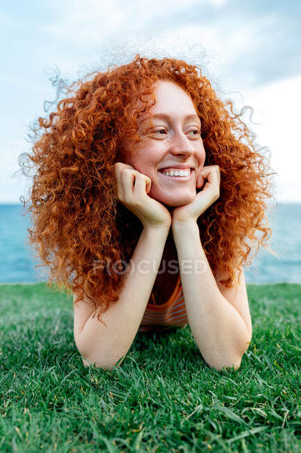 Glücklich gelockte rothaarige Frau mit Sommersprossen liegt auf dem Rasen und schaut weg auf die Küste des Meeres — Stockfoto