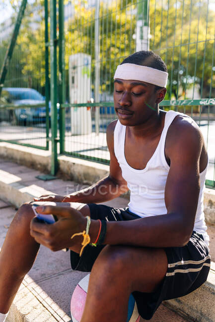 Afro-americano jogador de basquete masculino sentado em campo de esportes e smartphone de navegação no dia ensolarado no verão — Fotografia de Stock