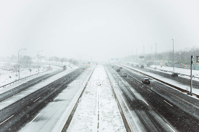 Coches conduciendo por una carretera de asfalto cubierta de nieve en un sombrío día de invierno durante las nevadas en Madrid - foto de stock
