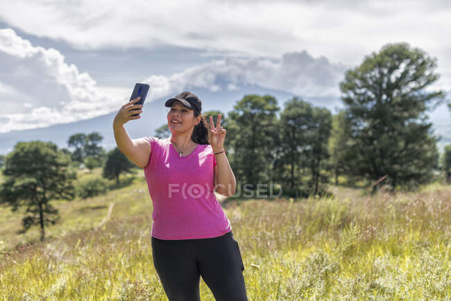 Женщина-туристка делает селфи на смартфоне, стоя на травянистом лугу с деревьями во время похода — стоковое фото