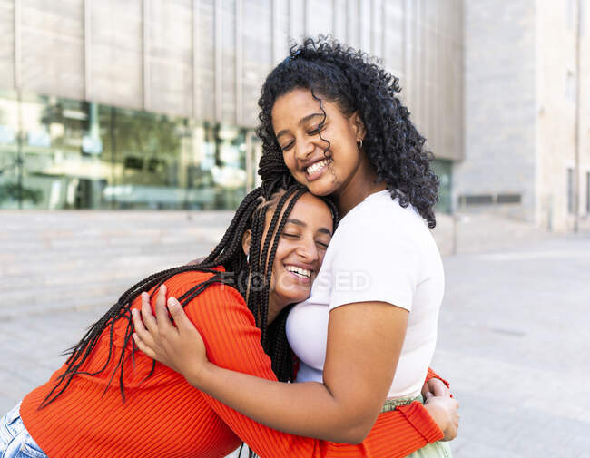 Namoradas multirraciais alegres em roupas da moda com olhos fechados e abraçando na rua da cidade durante o dia — Fotografia de Stock