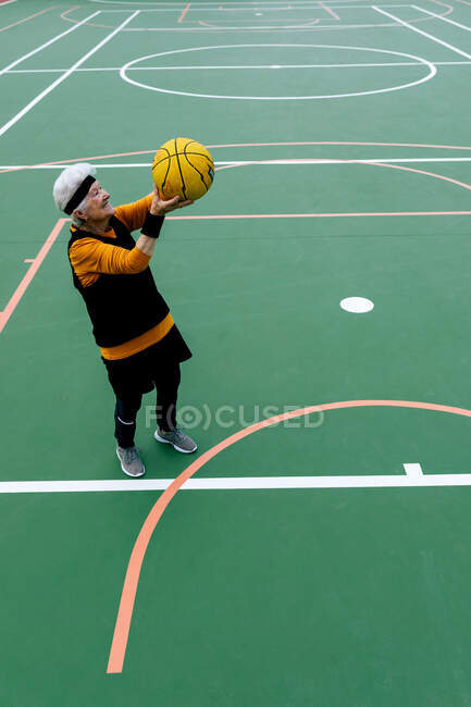 Старша зріла жінка в активному одязі кидає м'яч в обруч, граючи в баскетбол на громадському спортивному майданчику на вулиці — стокове фото
