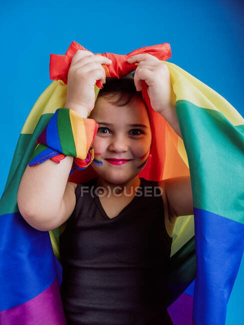 Petite fille avec un drapeau arc-en-ciel attaché sur la tête regardant la caméra tout en se tenant debout sur fond bleu — Photo de stock