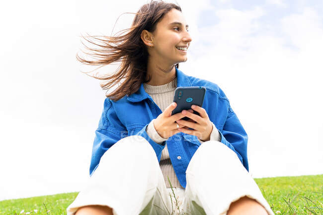 Low angle of glad female sitting on hill in nature on windy day and surfing Internet on mobile phone while looking away — Stock Photo
