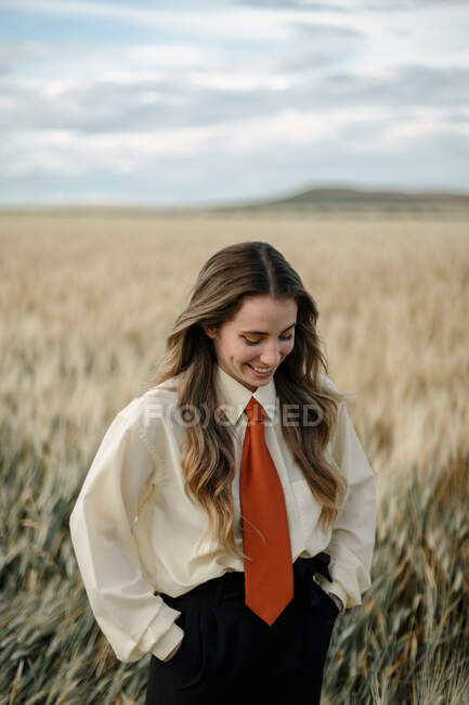 Позитивная молодая женщина в белой рубашке и красном галстуке, стоящая с руками позади пшеничных шипов в сельской местности — стоковое фото