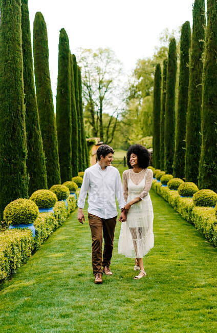 Fröhliches multiethnisches Paar hält Händchen und geht auf grünem Pfad in Gasse im Garten, während man sich gegenseitig ansieht — Stockfoto