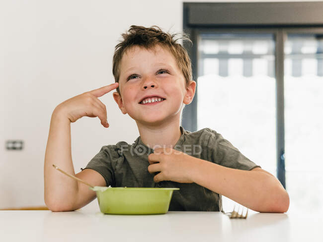 Porträt eines entzückenden, fröhlichen Jungen, der mittags zu Hause am Tisch sitzt und mit breitem Lächeln Gesten macht — Stockfoto