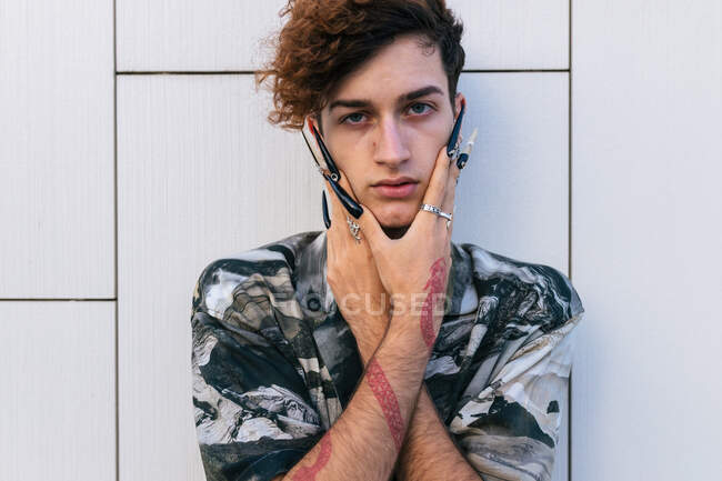 Hombre joven y vanidoso en ropa elegante con uñas largas de pie en la pared de baldosas mirando a la cámara - foto de stock