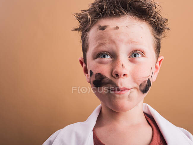 Grübelndes Kind mit blauen Augen und Farbklecksen im Gesicht, das auf beigem Hintergrund aufblickt — Stockfoto