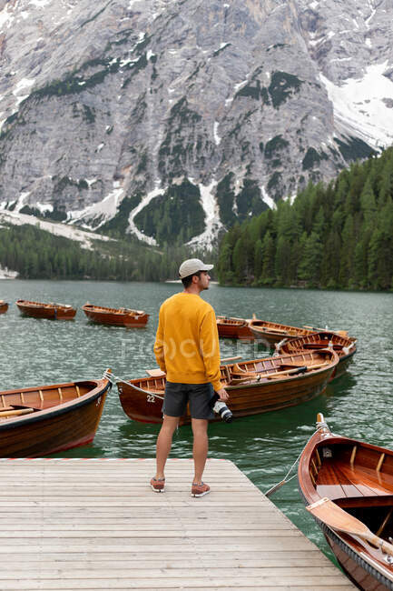 На задньому плані нерозпізнаний чоловік - дослідник фотограф з професійною фотокамерою в жовтому светрі стоїть на тлі озера Брейз з пришвартованими дерев 