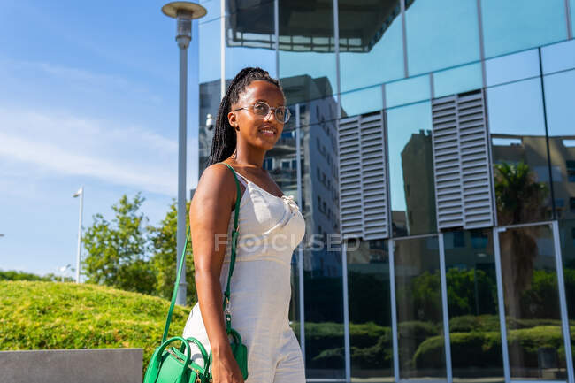 Femme afro-américaine élégante en robe d'été blanche et avec des tresses marchant le long de la rue dans la ville de Barcelone et détournant les yeux — Photo de stock
