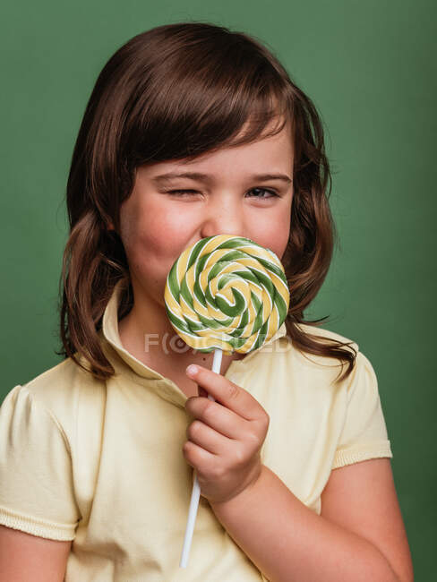 Divertido niño preadolescente lamiendo dulce remolino piruleta sobre fondo verde en el estudio y mirando a la cámara - foto de stock