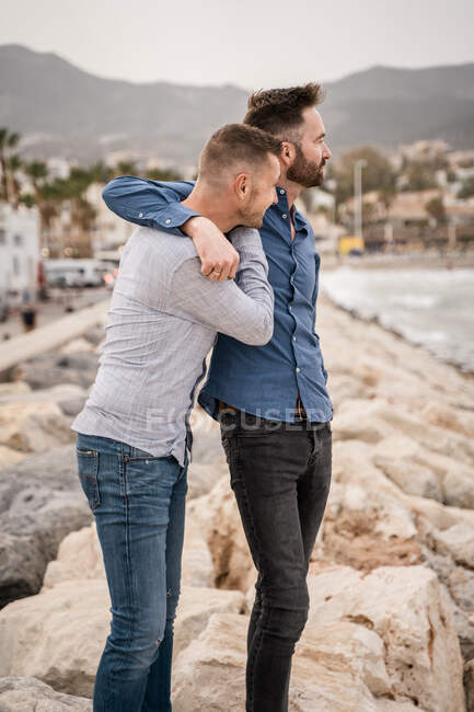 Casal de homens homossexuais em camisas e jeans abraçando enquanto olha para longe na costa rochosa contra o oceano e montanha — Fotografia de Stock