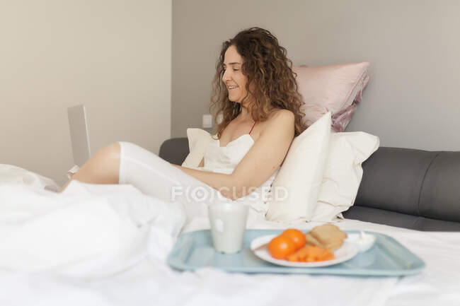 Vue latérale d'une femme d'affaires aux cheveux bouclés assise dans le lit et travaillant avec son ordinateur portable et un plateau avec déjeuner — Photo de stock