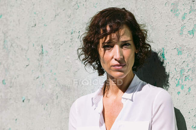 Thoughtful female wearing elegant clothes standing sunlight on shabby wall and looking at camera — Stock Photo