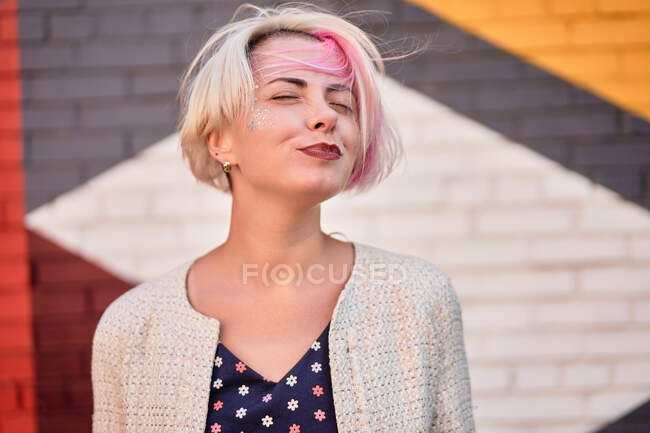 Happy fêmea alternativa no vestido da moda e com cabelo curto tingido de pé contra a parede de tijolo colorido na rua e olhos fechados — Fotografia de Stock