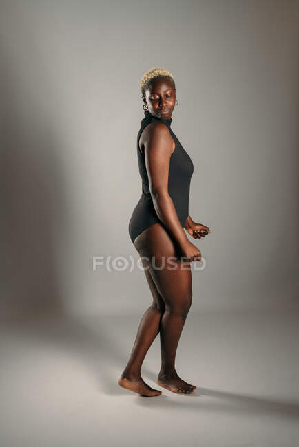 Side view of curvy African American female in bodysuit standing on gray background in studio for concept of body positivity — Stock Photo