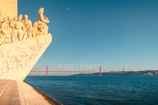 Famous Padrao dos Descobrimentos monument located on embankment of Tagus River against cloudless sundown sky and 25 de Abril Bridge in Lisbon, Portugal — Stock Photo
