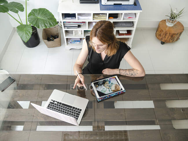 Desde arriba de diseñador gráfico femenino utilizando netbook y tableta con lápiz óptico mientras se trabaja en el proyecto en la mesa en el estudio creativo - foto de stock