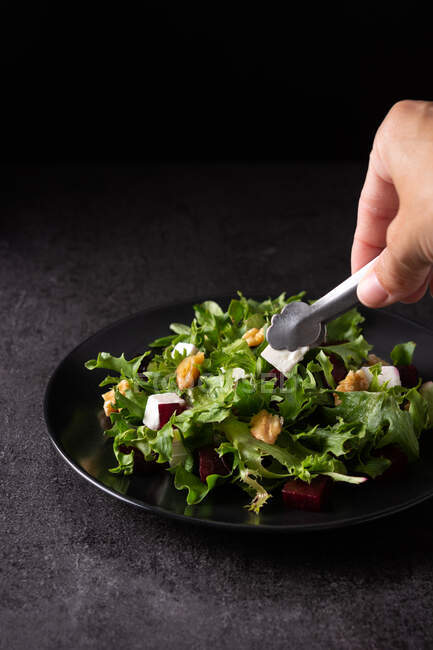 Crop anonymous cook with kitchen tongs serving appetizing beetroot salad with Tofu cheese and lettuce leaves on plate on dark table in studio — Stock Photo