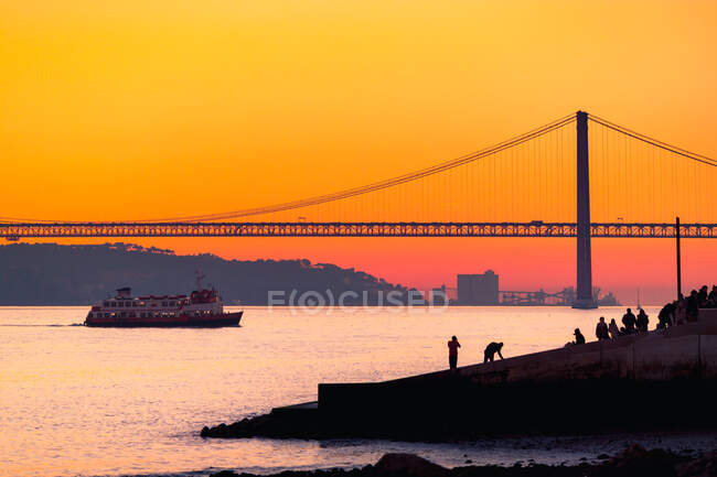 Набережні, розташовані біля річки Таг на поромі біля силуету 25 de Abril Bridge проти оранжевого сонячного неба в Лісабоні (Португалія). — стокове фото