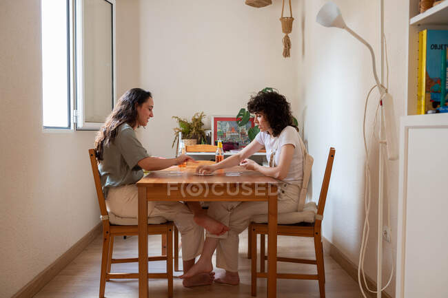 Vista laterale di una coppia di donne LGBT deliziate sedute a tavola e che giocano a carte durante il weekend a casa — Foto stock