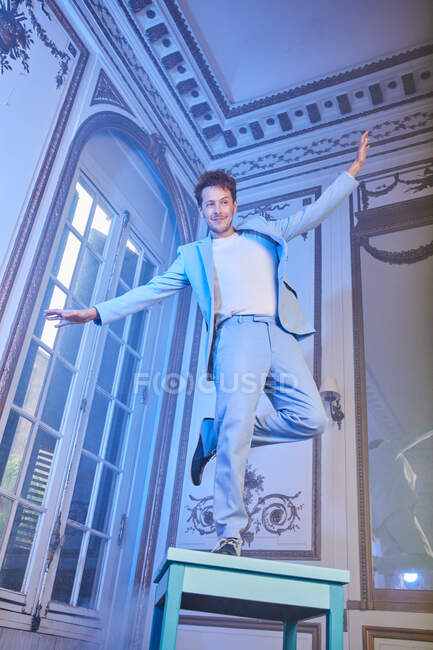 Знизу виразного щасливого чоловіка в стильному костюмі балансує на столі в похмурій кімнаті з блакитним неоновим світлом, дивлячись далеко — стокове фото