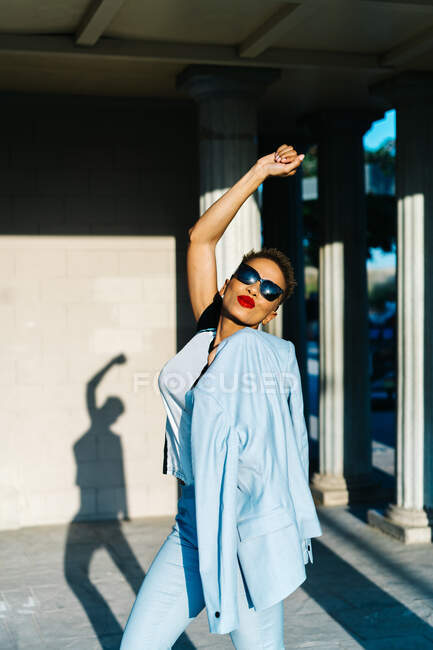 Вид збоку збудженої етнічної жінки в стильному одязі та сонцезахисних окулярах, що розважаються на міському тротуарі на сонячному світлі — стокове фото