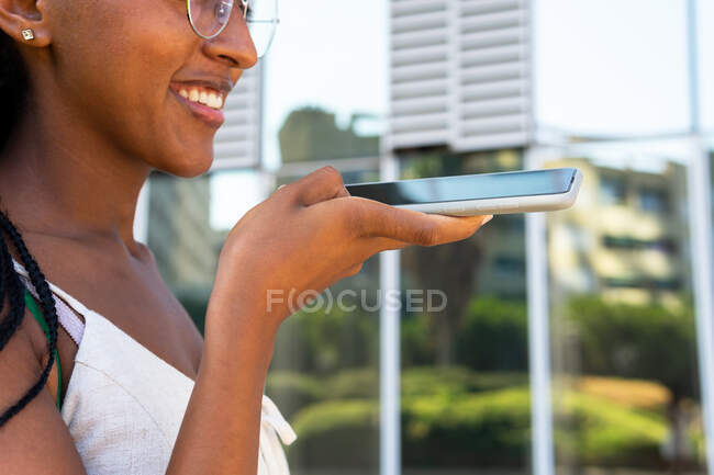 Seitenansicht einer lächelnden Afroamerikanerin, die eine Audiobotschaft auf ihrem Handy aufzeichnet, während sie im Sommer in Barcelona auf der Straße steht — Stockfoto