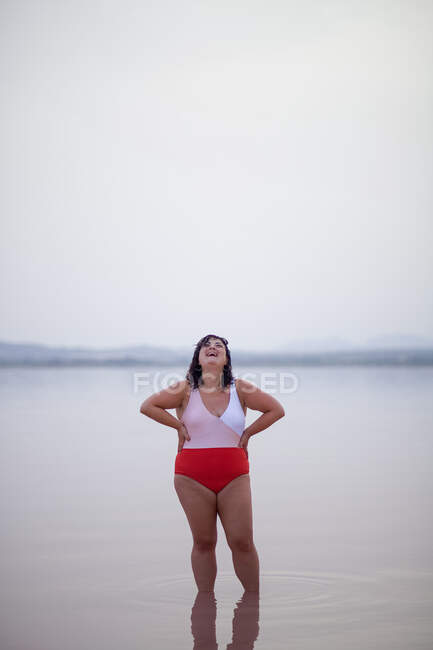 Positive curvy female in swimsuit standing with hands on waist on beach near pink pond and looking up while enjoying summer holiday — Stock Photo