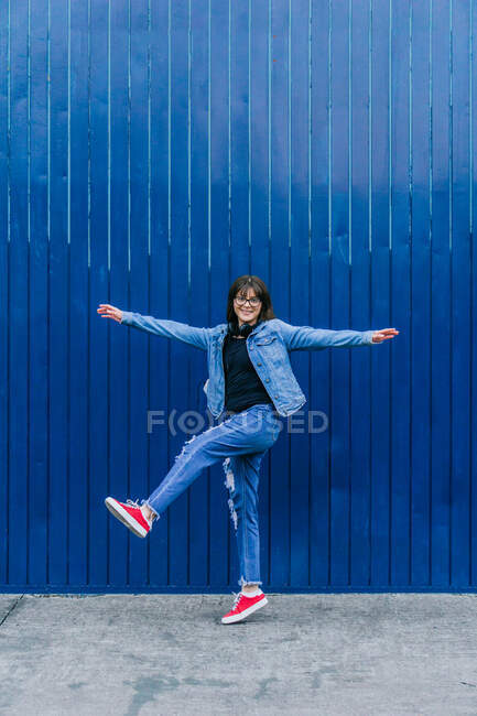 Emocionado hipster femenino en traje de mezclilla saltando con los brazos extendidos sobre fondo azul en la ciudad y mirando a la cámara - foto de stock