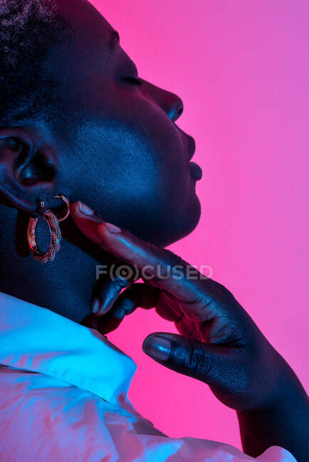 Вид сбоку на нежную афроамериканскую модель в тризубчатых серьгах, касающихся шеи, в студии с неоновым светом на розовом фоне — стоковое фото