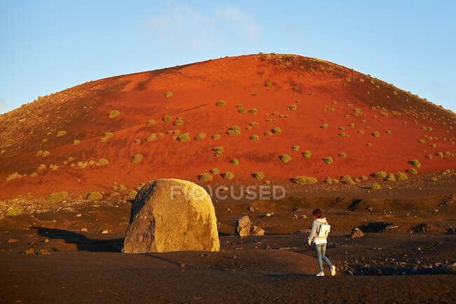 Voyageuse marchant près de rocher et colline sans eau couverte de buissons par une journée ensoleillée d'été à Fuerteventura, Espagne — Photo de stock