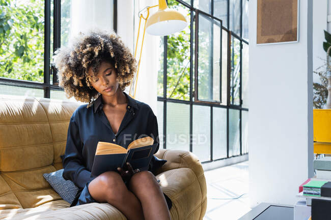 Художня афроамериканська жінка читає цікаву книжку, коли холоне на зручному дивані у вітальні і отримує задоволення від вихідних — стокове фото