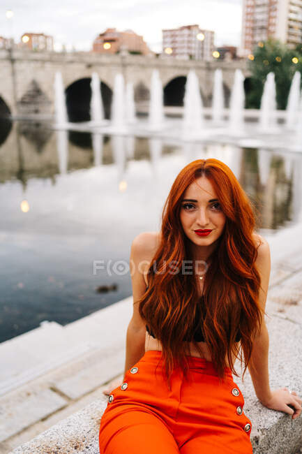 Jolie femelle avec de longs cheveux roux et un pantalon orange vif assis sur la frontière sur la promenade en ville regardant la caméra — Photo de stock