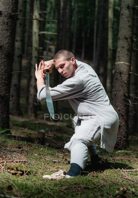 Hombre de cuerpo entero con ropa tradicional practicando la postura de la espada durante el entrenamiento de kung fu en el bosque - foto de stock